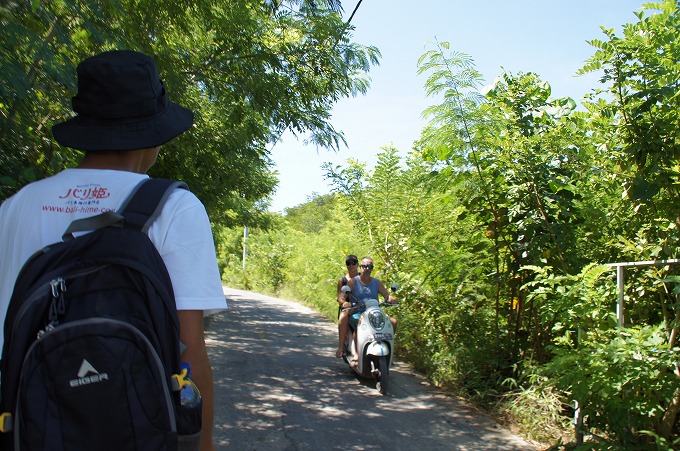 レンボンガン島の道を歩くスタパとバイクに二人乗りをする観光客