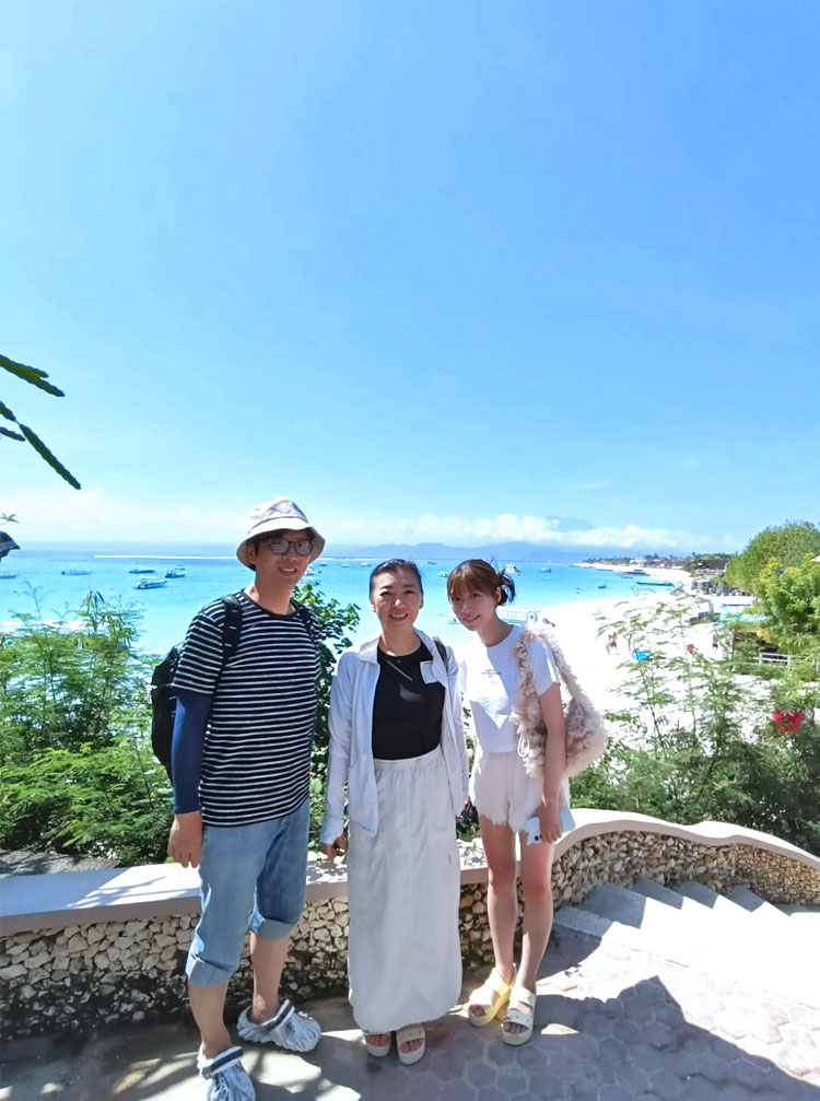 レンボンガン島の海を一望できる高台より青い空とエメラルドグリーンの海とホワイトサンドを背に笑顔のご夫婦と娘様の3人ご顔族様